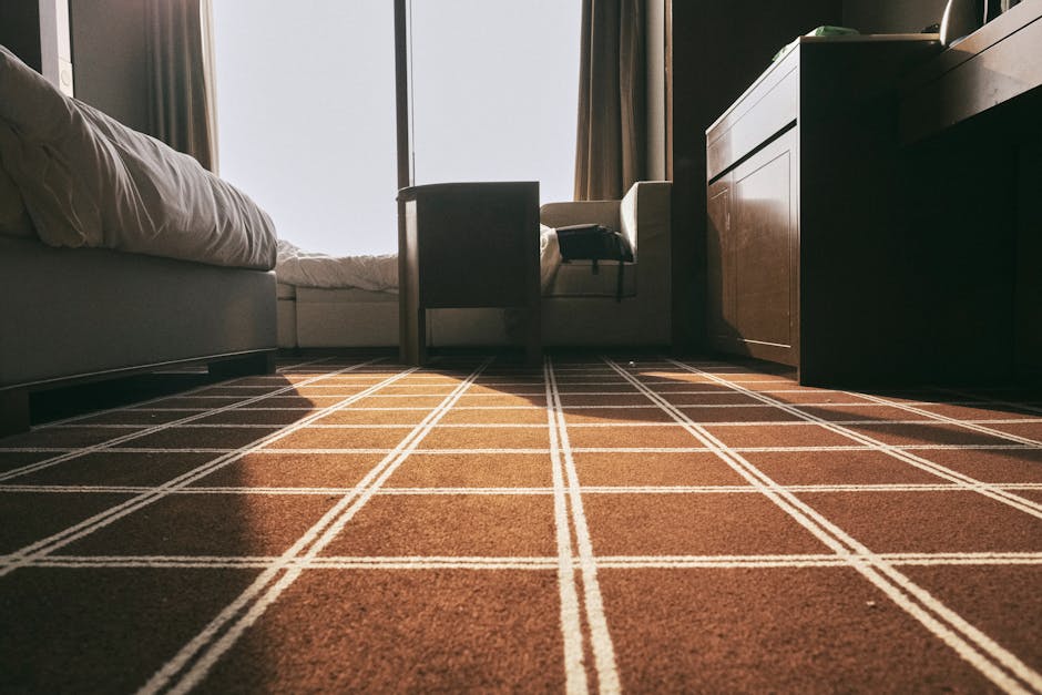Close-up of a person's hands using a cloth to clean the interior of a yellow and black vacuum cleaner, placed on a patterned rug in a domestic setting. The vacuum cleaner's open top reveals the dust c