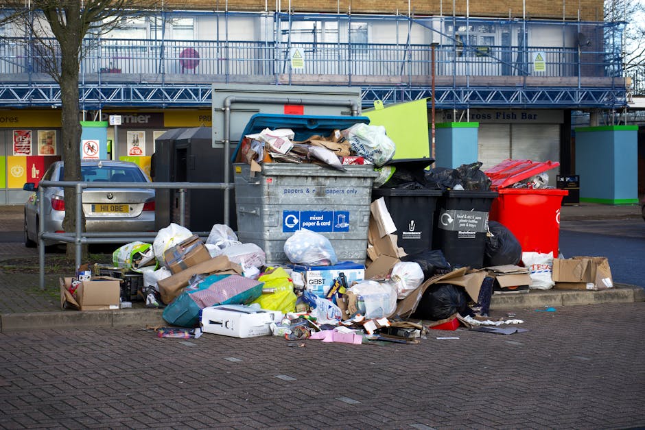 Piles of mixed household waste including cardboard boxes, plastic bags, paper clutter, and black bin liners are overflowing from a large recycling bin situated on a paved sidewalk in front of a commer