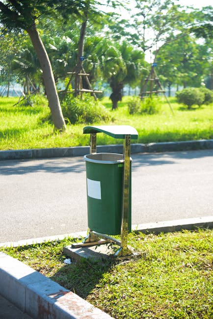 Piles of mixed household waste including cardboard boxes, plastic bags, paper clutter, and black bin liners are overflowing from a large recycling bin situated on a paved sidewalk in front of a commer