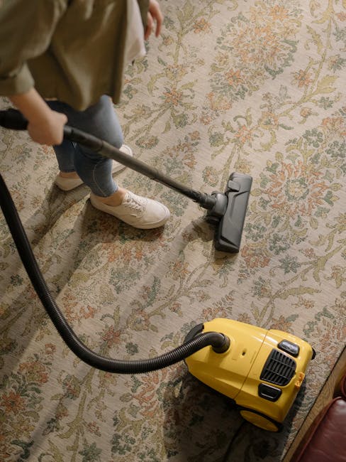 A person using a yellow and black vacuum cleaner to perform surface cleaning on a patterned carpet in a residential setting. The individual is kneeling on the carpet, placing a white cloth or filter i